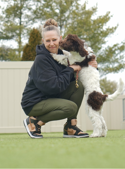 Canine caretaker kneeling next to and getting hugs and kissed from a brown and white doodle puppy in the backyard of Greenleaf Pet Resort.